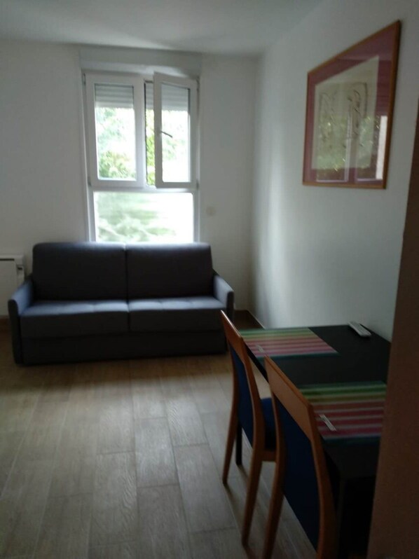 A room with a sofa, a small table, and a chair. There is a window with blinds, and a framed picture on the wall. - Intimiste Studio - 2P - Malakoff (Malakoff)