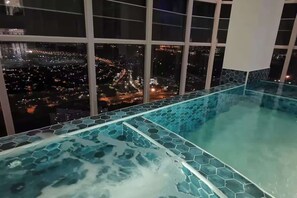 An indoor infinity pool with a hexagonal tile design, large windows offering a cityscape view at night. - Cozy 1BR Suite at TriTower Steps to JB CIQ (Johor Bahru)