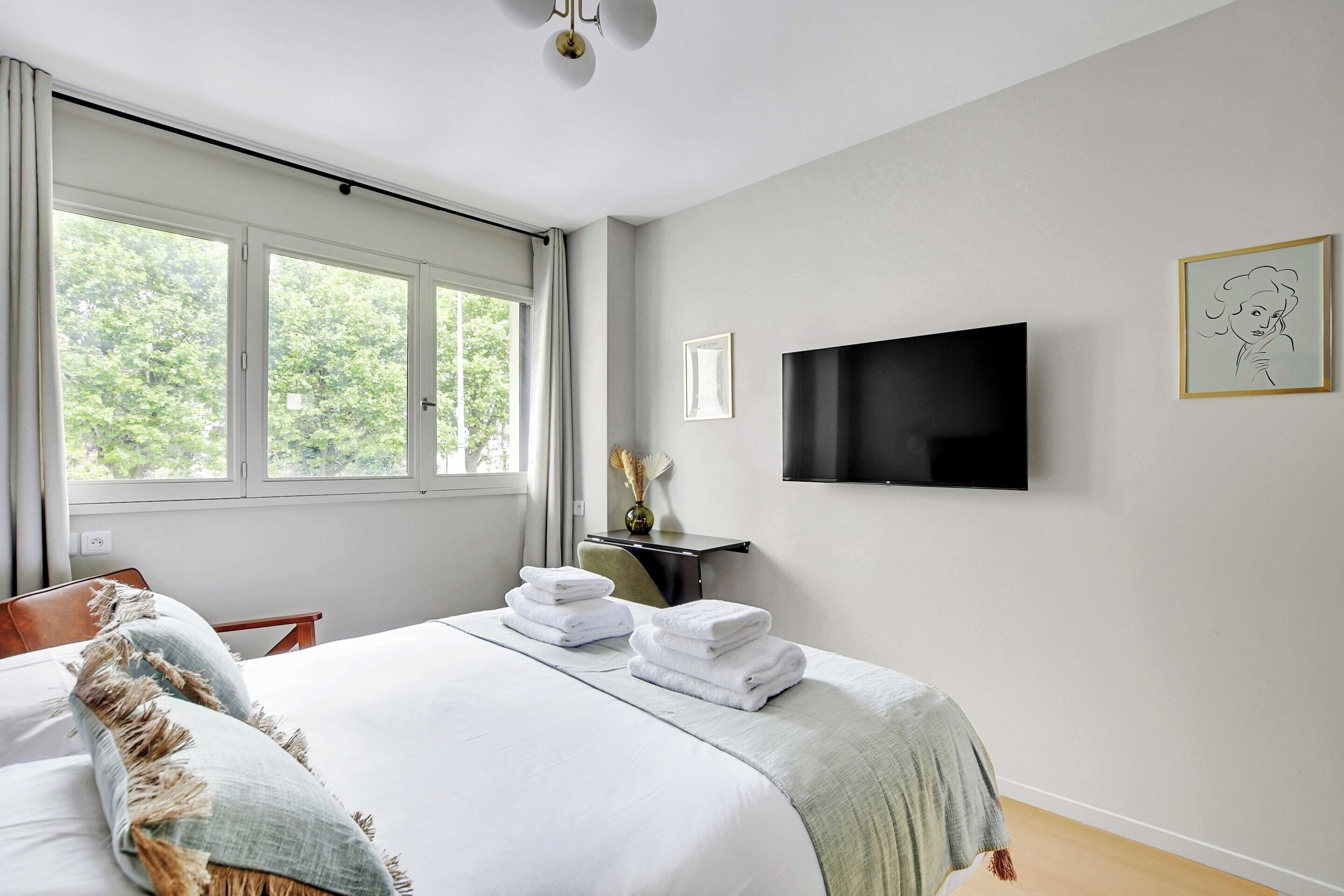 A neatly made bed with folded towels, a flat-screen TV mounted on the wall, and a window with curtains.