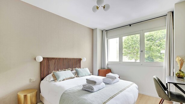Una moderna camera d'albergo con un letto grande, una testiera in legno, un comodino con un vaso e vista sugli alberi dalla finestra.