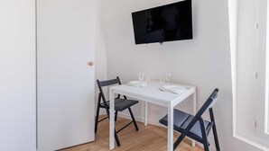 Un petit coin repas, tout en blanc, avec une table dressée pour deux personnes, deux chaises noires et un téléviseur à écran plat fixé au mur.