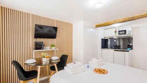 Quarto de hotel com parede revestida de painéis de madeira, televisão de tela plana, mesa pequena com cadeiras, cama com lençóis brancos e área de cozinha compacta.