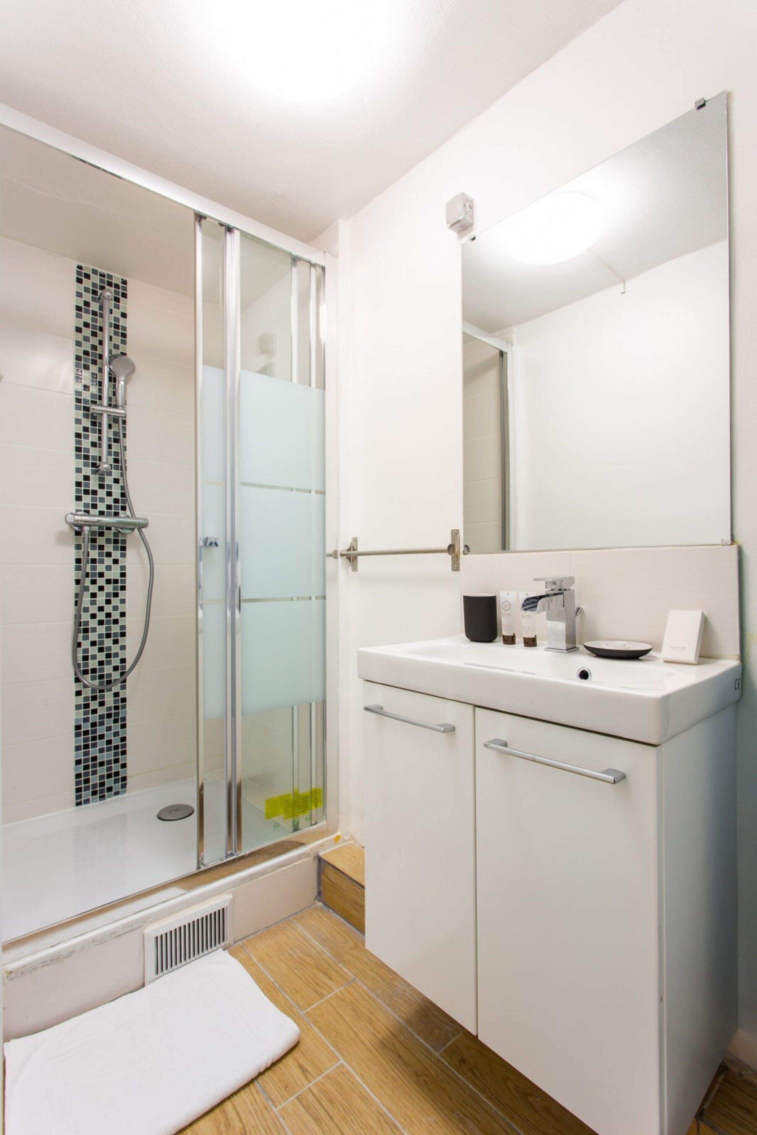 A modern bathroom with a glass shower enclosure, a white sink with a mirror above it, and a wooden floor.