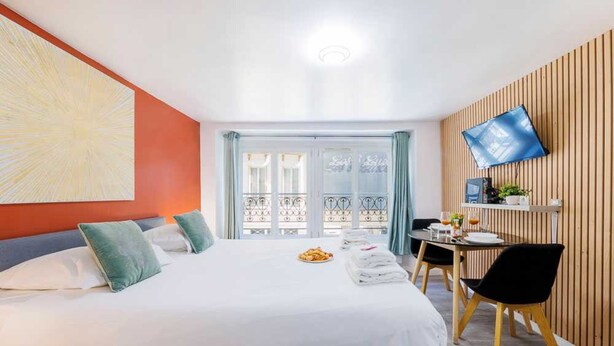 Quarto de hotel com uma cama grande, uma escrivaninha com cadeira, uma TV fixada na parede e uma janela com cortinas.