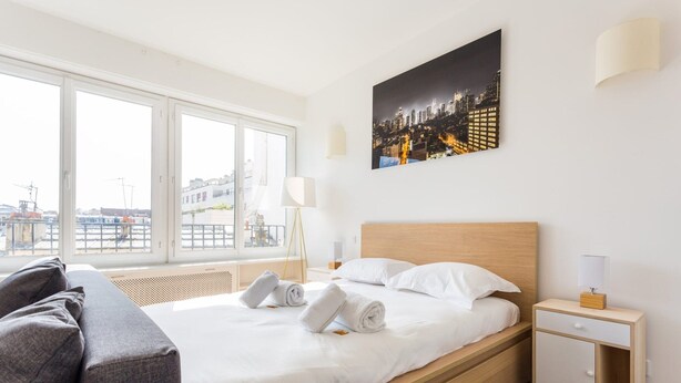 A modern hotel room with a large bed, a nightstand, a lamp, and a cityscape view through large windows. - Superbe Studio - 4P - Trocadero (Paris)