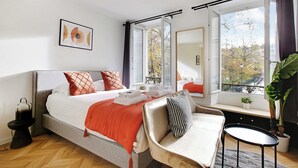 A modern bedroom with a large bed, a bedside table, a chair, a window with curtains, and a potted plant. - Welcoming Studio for 2 People - Bastille (Paris)
