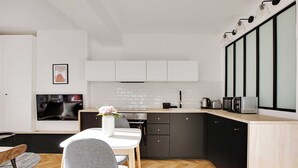 A modern kitchen with a dining area, featuring a herringbone wood floor, white cabinetry, and a black countertop. - Welcoming Studio for 2 People - Bastille (Paris)