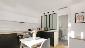 A modern kitchen with a white countertop, black cabinets, and a dining area with a white table and grey chairs. - Welcoming Studio for 2 People - Bastille (Paris)