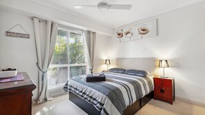 A bedroom with a bed, bedside lamps, a nightstand, a window with curtains, a ceiling fan, and a wall art piece. - Esplanade Beachfront at Victor- 2 - 12A Esplanade, (Victor Harbor)
