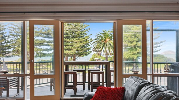 Ein Zimmer mit einer Schiebetür, die auf einen Balkon mit Blick auf Bäume und Gewässer führt. Es gibt ein schwarzes Ledersofa mit rotem Kissen und einen kleinen Tisch mit Stühlen.