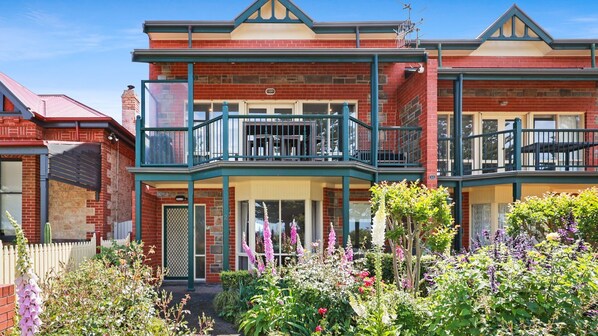 A two-story brick house with a balcony, a picket fence, and a garden with various flowers. - Esplanade Beachfront at Victor- 2 - 12A Esplanade, (Victor Harbor)