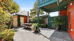 A patio with a table and chairs, surrounded by potted plants and a green fence. - Esplanade Beachfront at Victor- 2 - 12A Esplanade, (Victor Harbor)