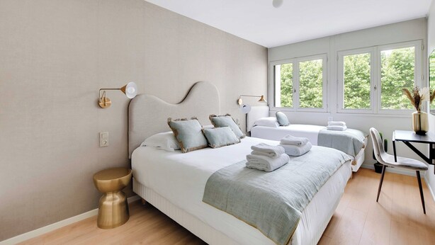 Una camera d'albergo moderna con un letto grande, un letto più piccolo, una scrivania e una sedia. Diverse finestre offrono luce naturale e vista sul verde.