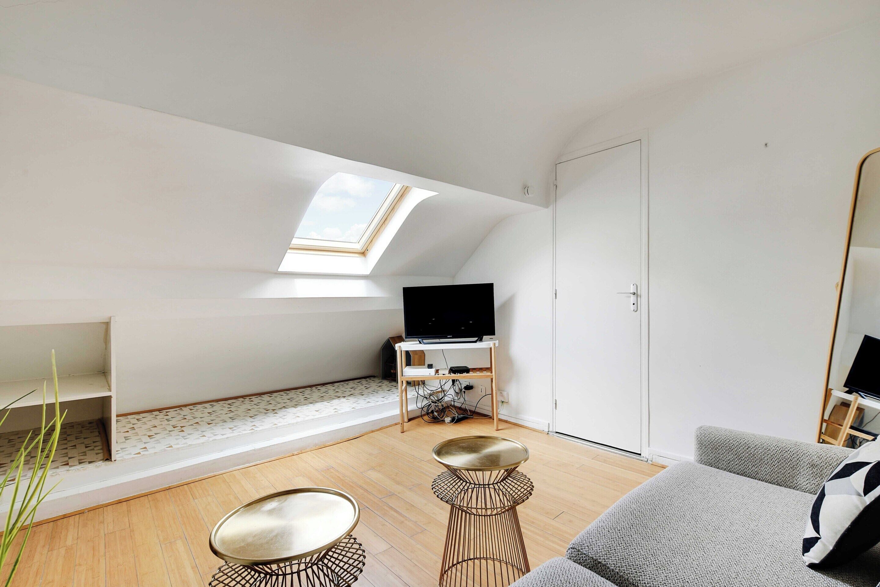 A modern loft bedroom with a skylight, a TV on a wooden stand, a grey sofa, and a small round table.