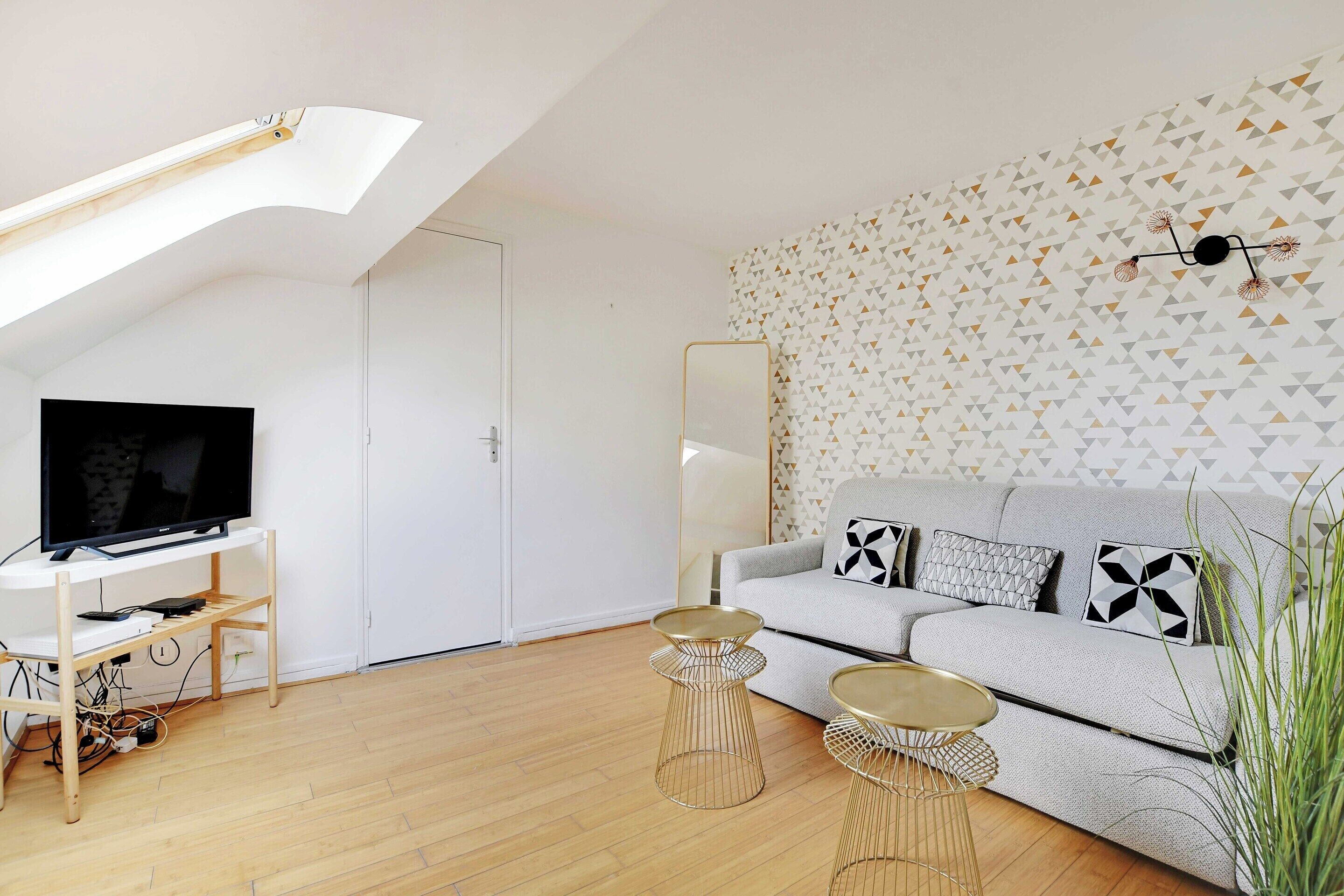 Ein modernes Wohnzimmer mit einem Fernseher auf einem Holzboden, einem grauen Sofa mit gemusterten Kissen und einer Wand mit geometrischem Muster.
