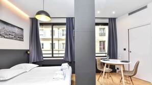 Una habitación de hotel moderna con una cama, una mesa de comedor con sillas y vista a un paisaje urbano a través de la ventana.