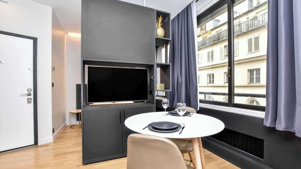 Una habitación moderna con una televisión de pantalla plana, una mesa redonda de comedor con sillas y un ventanal con vistas a un edificio.