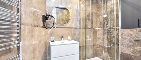 Un baño moderno con ducha envuelta en vidrio, un tocador blanco con espejo redondo y un perchero.