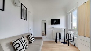 A modern living room with a sofa, two black and white patterned pillows, a mounted TV, a radiator, and a window with a view. - Cosy Studio for 2 People - Malakoff (Malakoff)