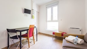 Ein kleiner, gut beleuchteter Raum mit Schreibtisch, zwei Stühlen, einer Couch, einem Fenster und einem an der Wand befestigten Fernseher.