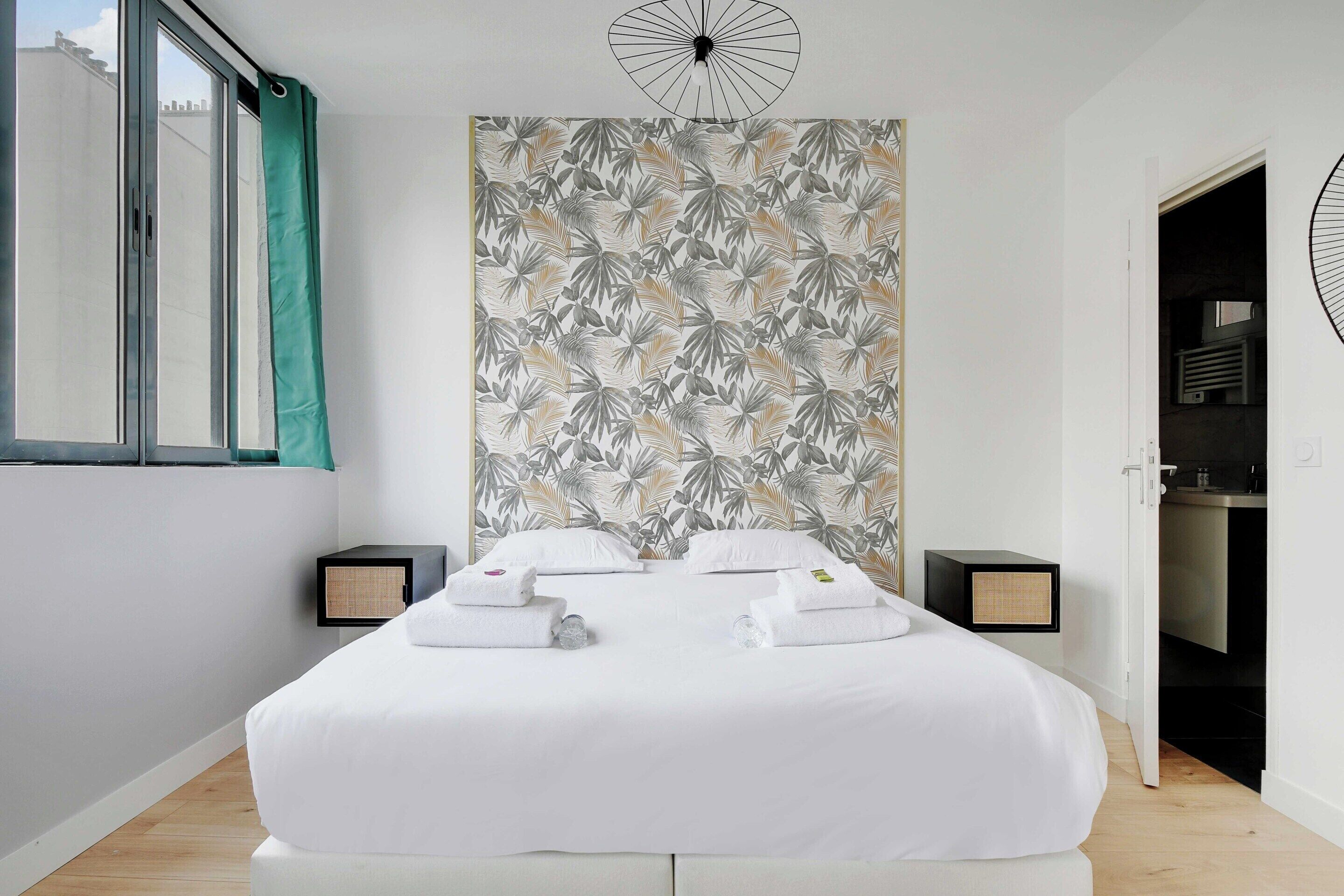 Habitación de hotel con una cama grande, dos mesitas de noche, una pared con diseño de hojas y una ventana con cortina.