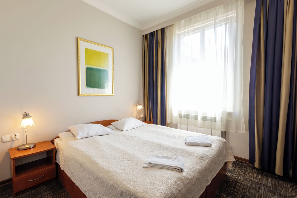Un cuarto de hotel con una cama bien tendida, una lámpara de noche, un cuadro en la pared y una ventana con cortinas.