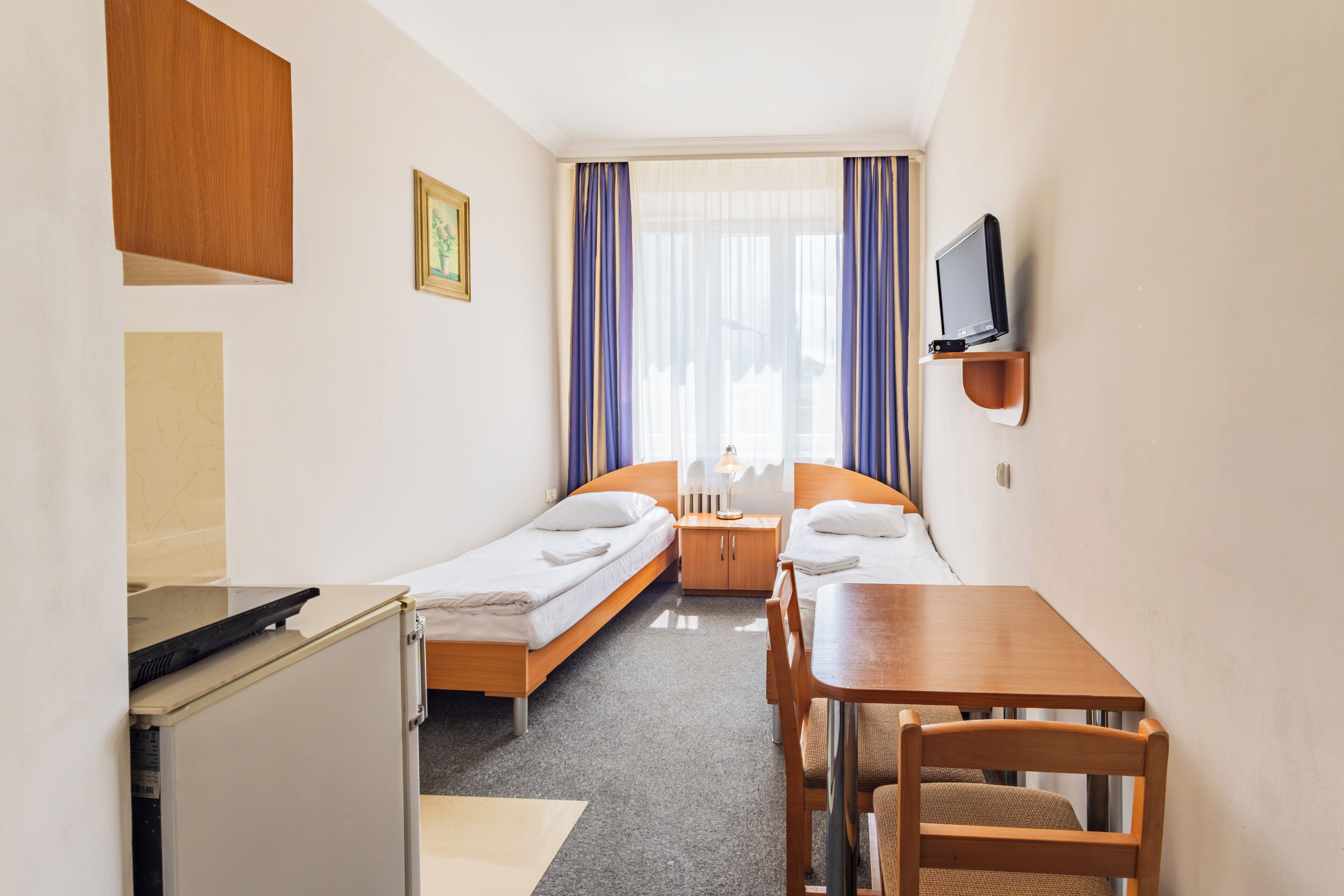 A compact hotel room with two single beds, a small table, a chair, a TV mounted on the wall, and a kitchenette area.