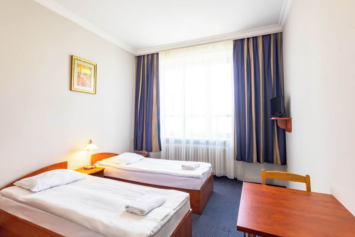 Habitación de hotel con dos camas individuales, un escritorio, una silla, un televisor sobre un mueble bajo y una ventana con cortinas.
