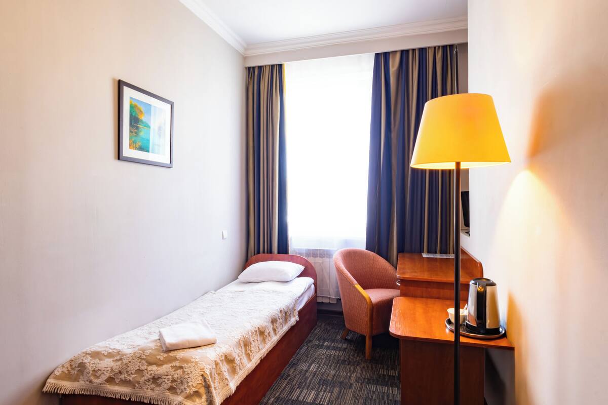 Una habitación de hotel pequeña con una cama individual, un escritorio con lámpara, una silla y un cuadro en la pared.