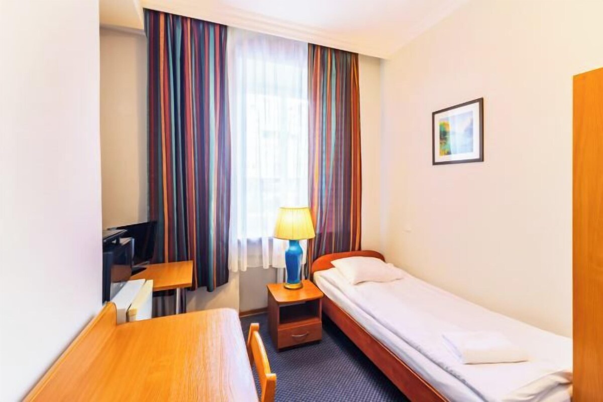 A compact hotel room with a single bed, a desk, a chair, a small table, a lamp, and a window with curtains.