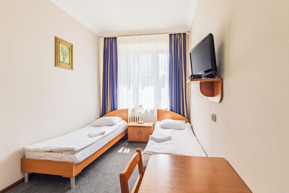 Una habitación de hotel pequeña con dos camas individuales, una mesa de madera, un televisor en la pared y una ventana con cortinas.