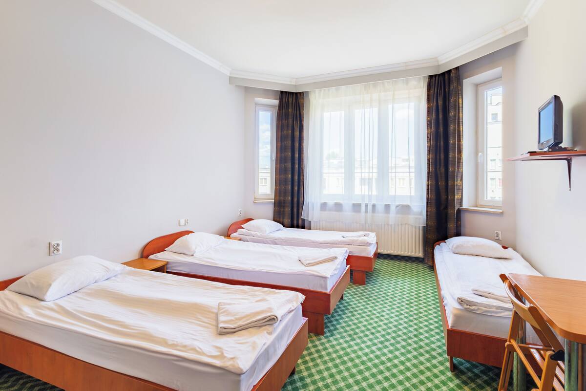 Habitación de hotel con tres camas individuales, una mesita, una silla, un televisor sobre una estantería y una ventana con cortinas.