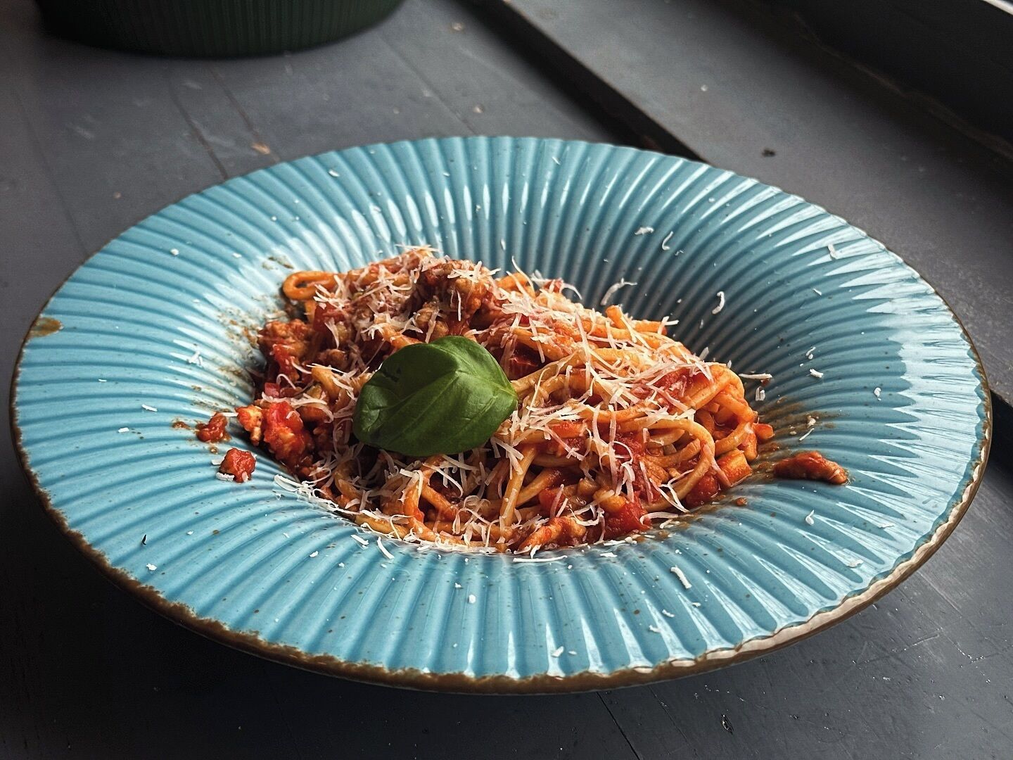 Un plato de espagueti con salsa de tomate y queso rallado, adornado con una hoja de albahaca, sobre un plato de cerámica azul.