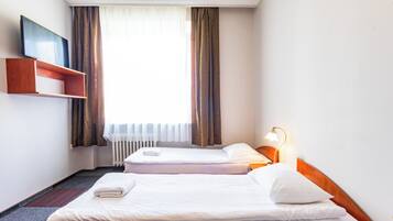 Habitación de hotel con dos camas, una mesita de noche con lámpara, un estante sobre las camas y una ventana con cortinas.