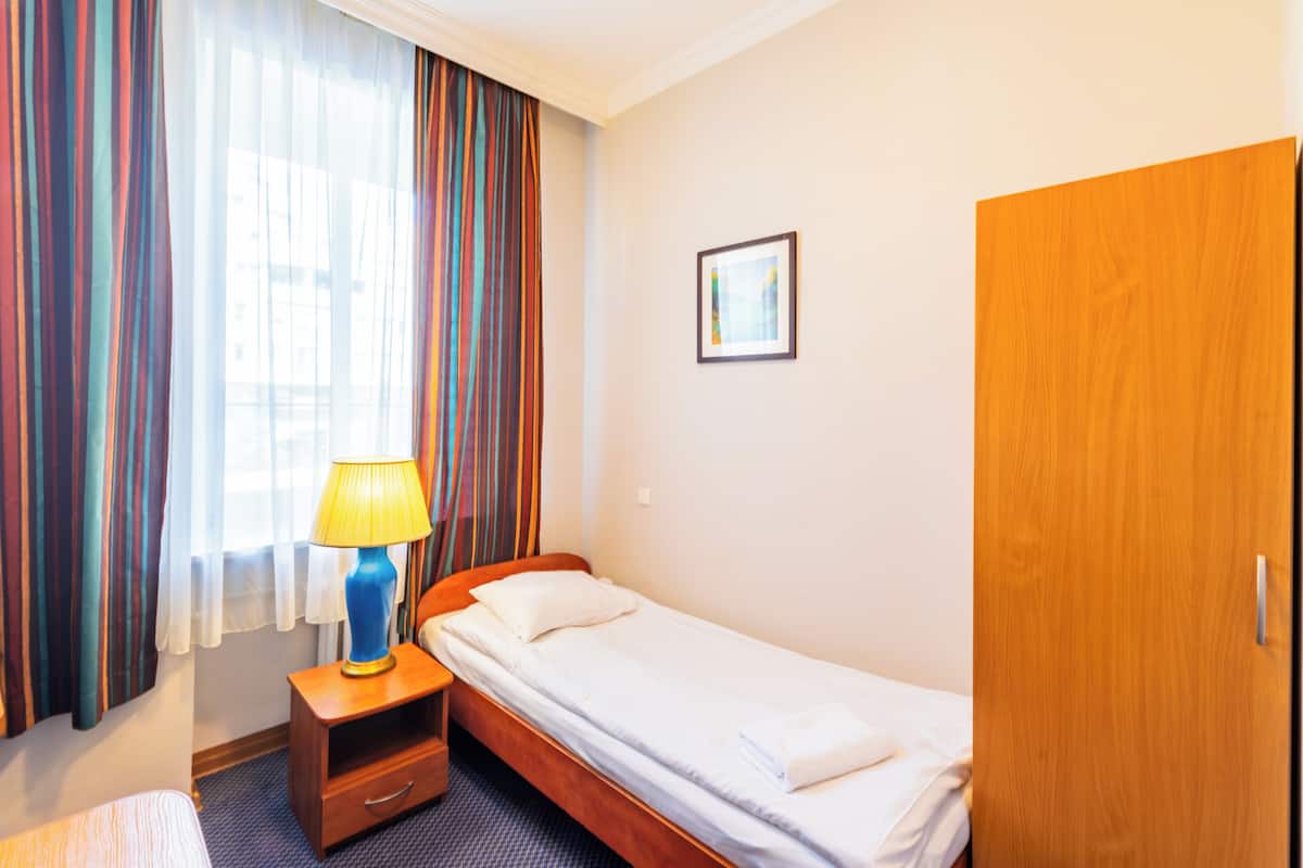 Habitación de hotel con una cama individual, una mesita de noche de madera con lámpara, armario y ventana con cortinas.