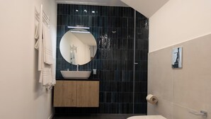 Et moderne badeværelse med en hvid vask, mørke flisevægge, et rundt spejl og et toilet.