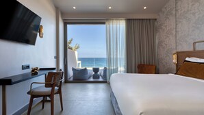 Habitación de hotel con una cama grande, un escritorio con silla, televisor montado en la pared y vista al mar a través de un ventanal amplio.