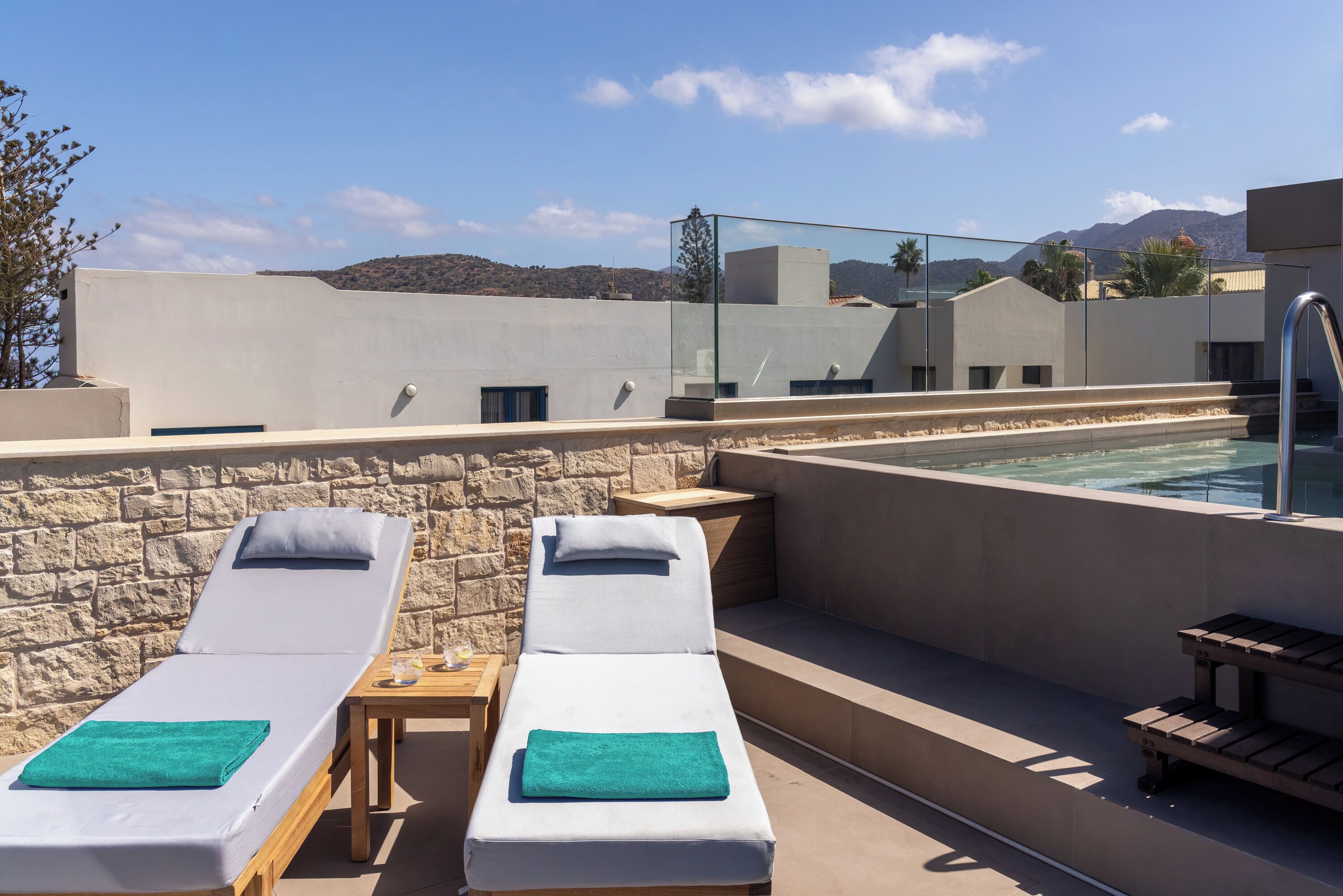 A rooftop pool area with two sun loungers, a stone wall, and a clear view of the mountains.