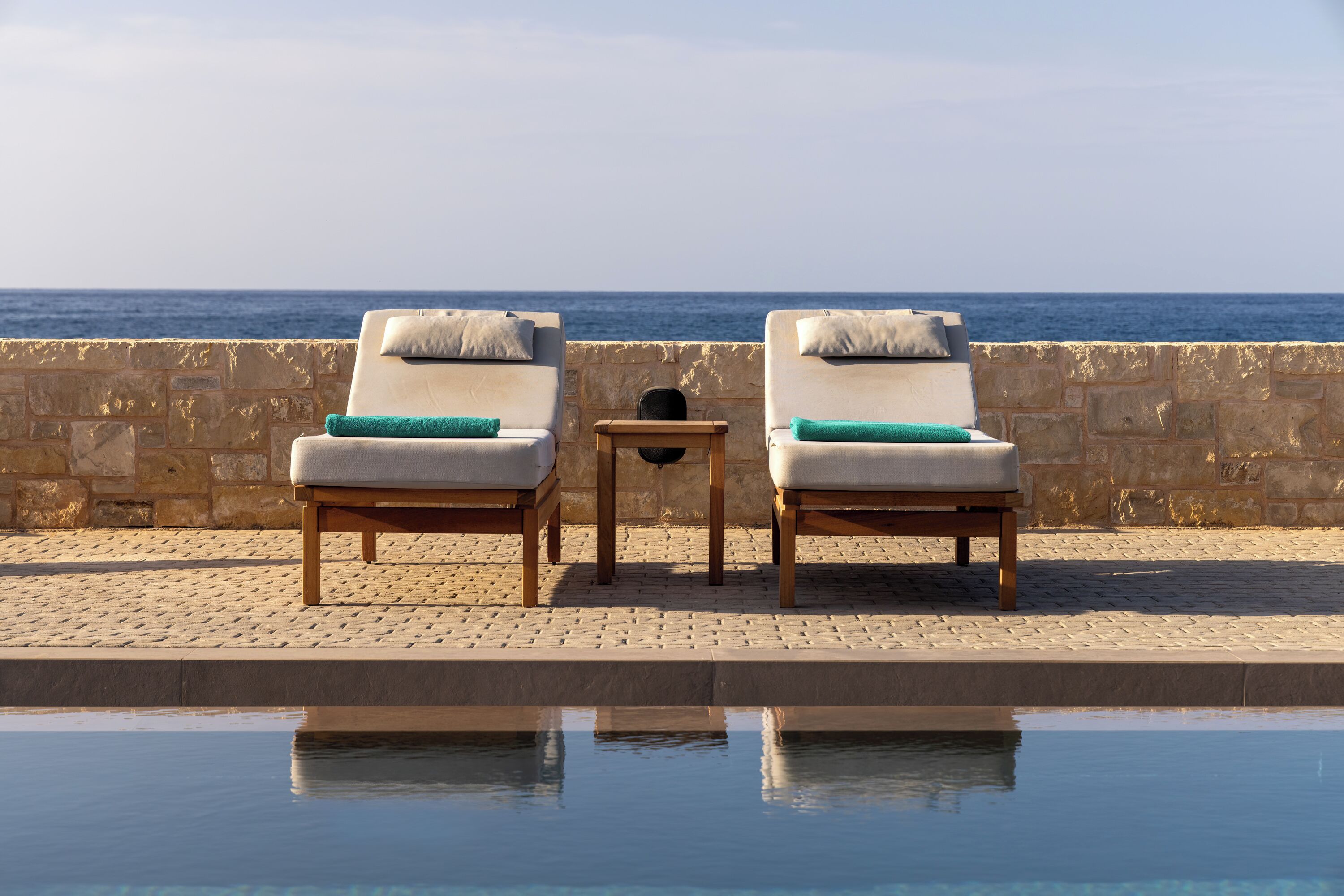 Due poltrone con cuscini accanto a una piscina, un muro in pietra e vista sul mare.