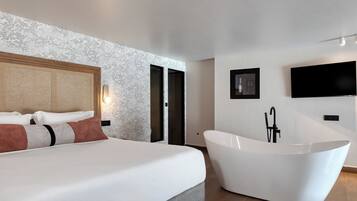 A modern bedroom with a large bed, a freestanding bathtub, and a flat-screen TV mounted on the wall.