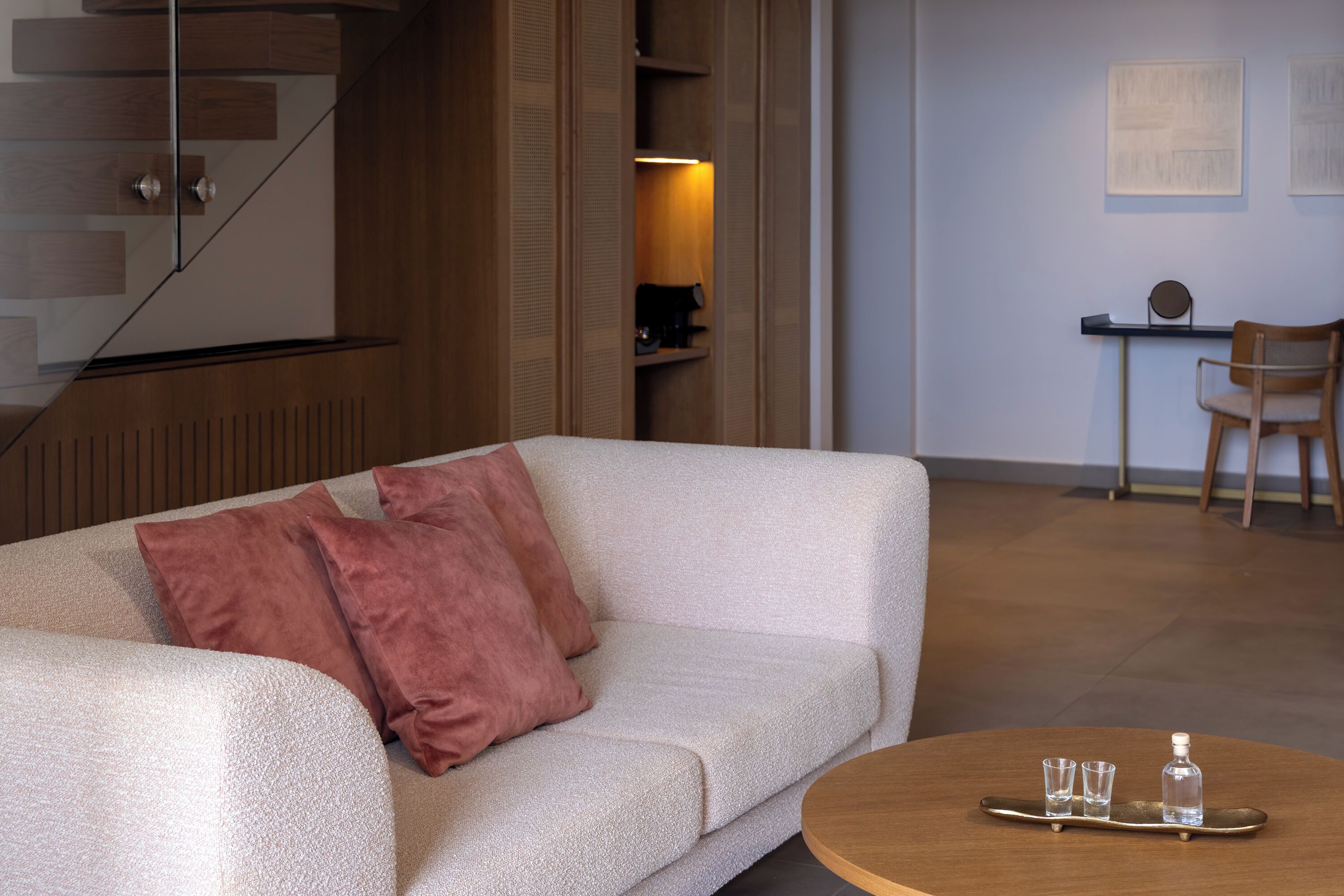 A modern living room with a sofa, a coffee table with glasses and a bottle, a wooden staircase, and a desk with a chair.