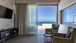 Habitación de hotel moderna con un gran ventanal que ofrece vista al mar, un televisor de pantalla plana montado en la pared, una consola oscura y dos sillones verdes.