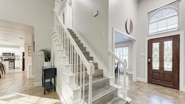 A spacious interior with a wooden staircase, a kitchen in the background, and a large wooden door with a glass panel. - 2 Story Home in Round Rock Sleeps 16 Guests (Round Rock)