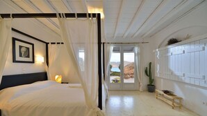 Ein Himmelbett mit Baldachin, ein gerahmtes Bild an der Wand, eine Nachttischlampe, eine Kakteenpflanze und eine hölzerne Bank mit einer Obstschale.