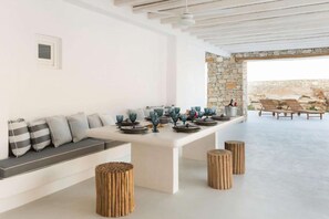 Un comedor moderno con una mesa blanca dispuesta para seis personas, una pared de piedra y vista al mar.