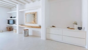 A minimalist room with white walls, a wooden ceiling, a large mirror, a TV on a shelf, a white cabinet, and a small trash bin. - Villa Conza 5 bed in Mykonos (Mykonos)