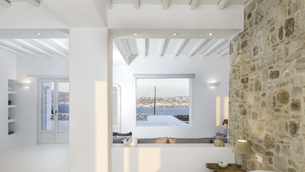 A modern bedroom with a stone wall, a large window with a view, a wooden nightstand, and a bed. - Villa Conza 5 bed in Mykonos (Mykonos)