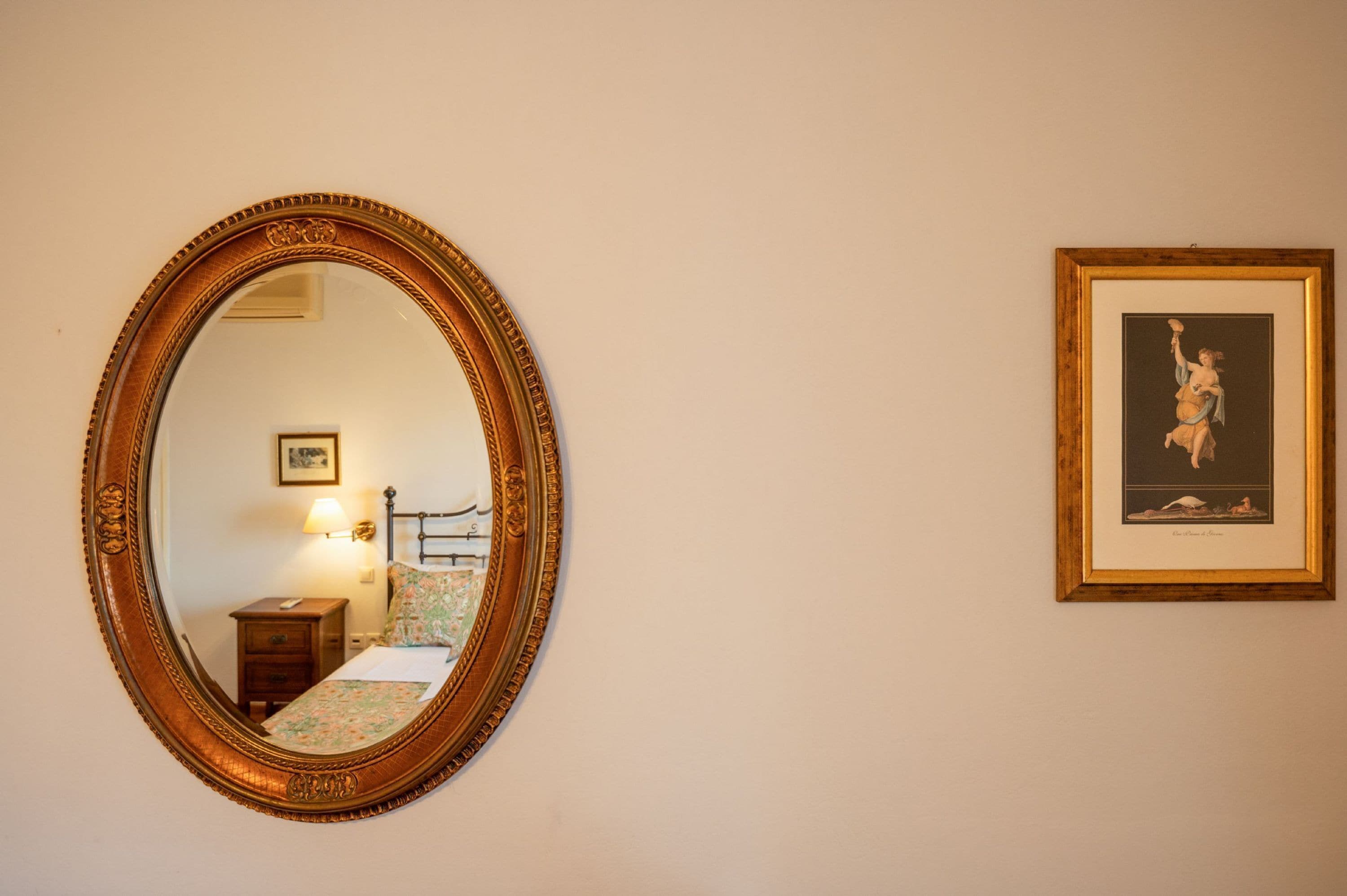 Une pièce comprenant un grand miroir décoratif, un tableau encadré accroché au mur, une table de chevet avec une lampe et un lit recouvert d’une literie à motifs floraux.