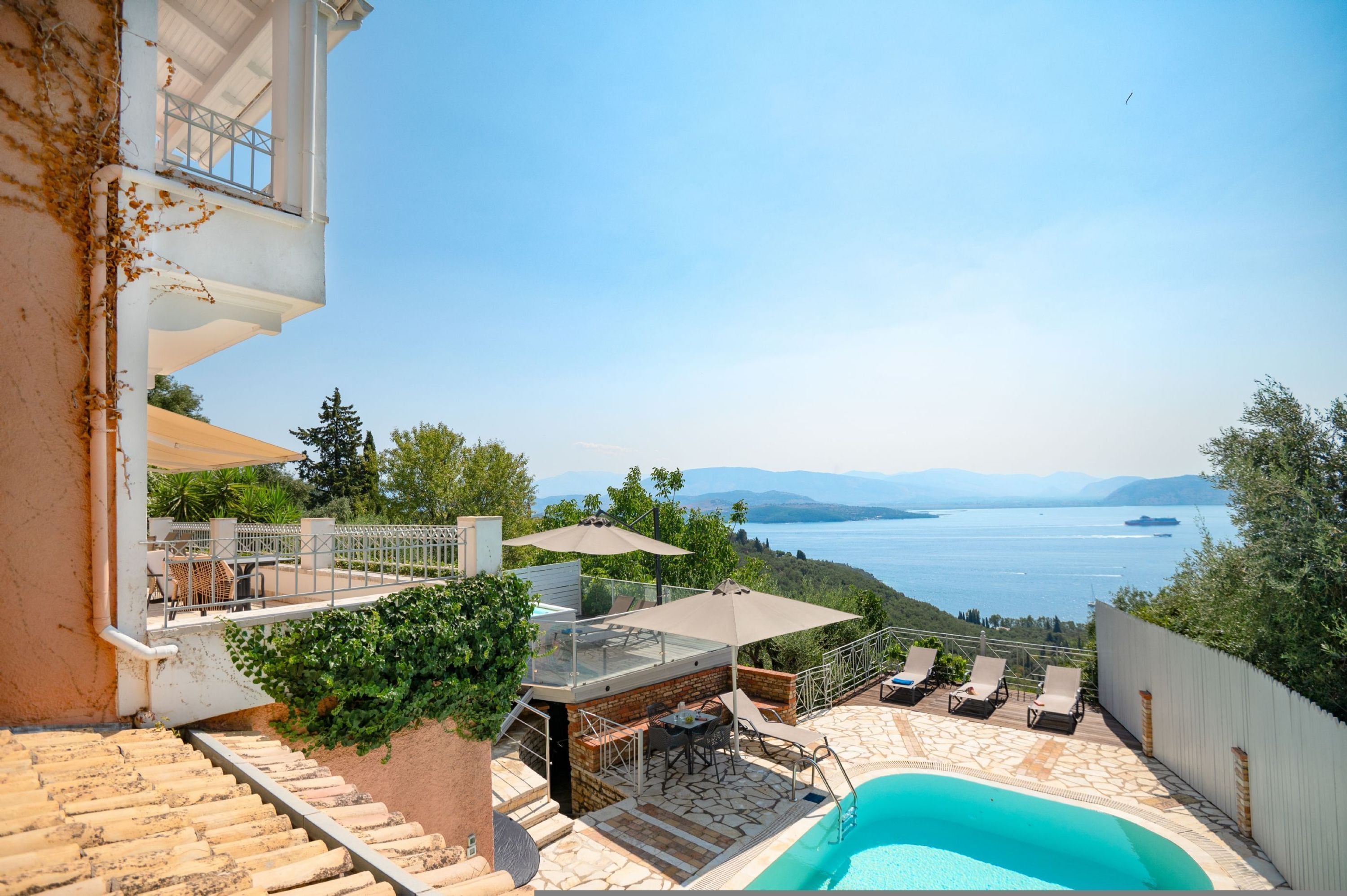 A pool area with a view of the sea, outdoor furniture, and a staircase leading to a balcony.