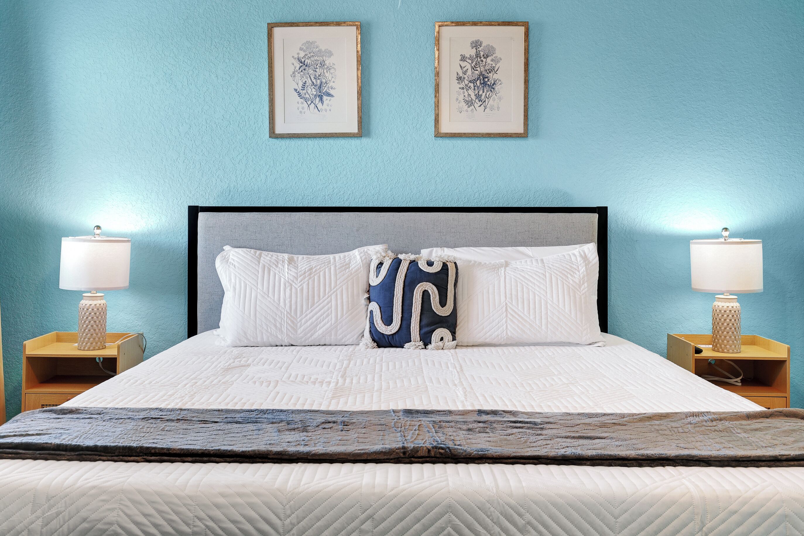 A neatly made bed with a patterned bedspread, two wall-mounted floral artwork, and bedside lamps on wooden nightstands.